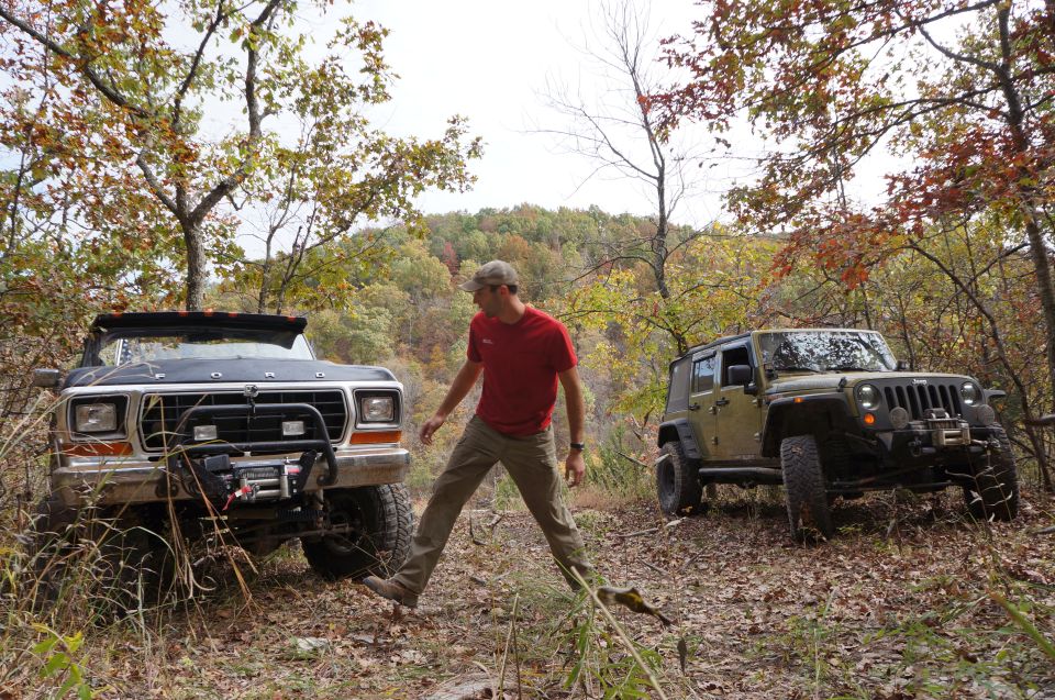 Man standing between a 1979 Ford Bronco and a Jeep Wrangler in a wooded area during fall
