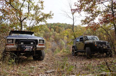 1979 Ford Bronco - Two off-road vehicles, a Ford Bronco and a Jeep Wrangler, are parked on a dirt path surrounded by autumn foliage.