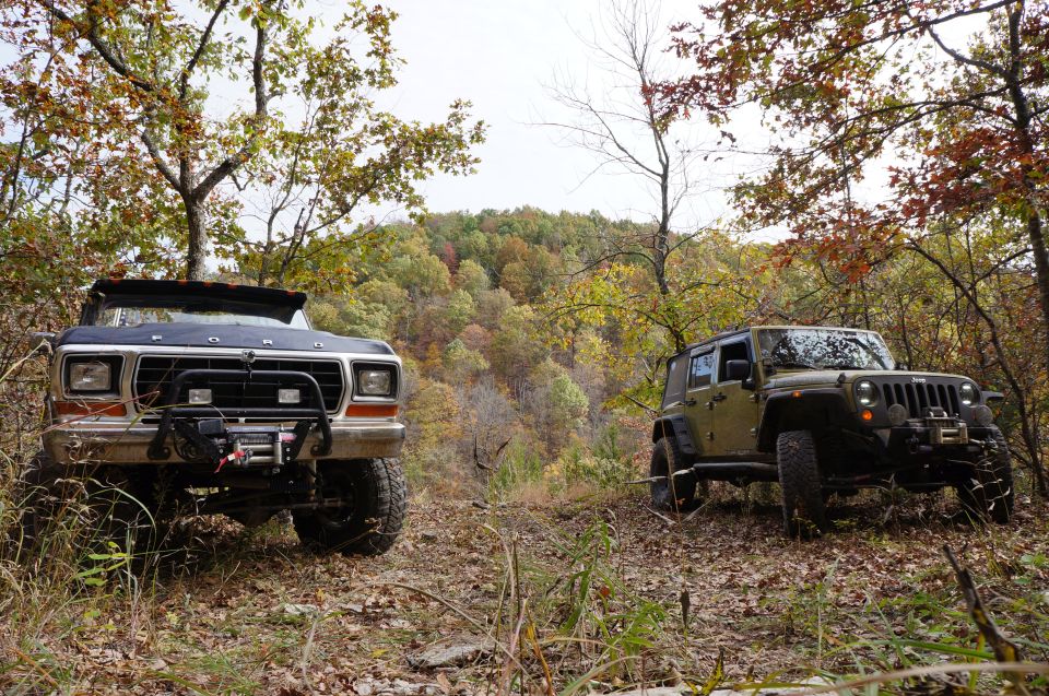 Ford Bronco front view, dark color, off-road modifications, parked on dirt path with autumn trees