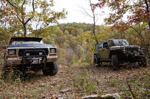 1979 Ford Bronco - Two off-road vehicles, a Ford Bronco and a Jeep Wrangler, are parked on a leaf-covered trail during autumn.
