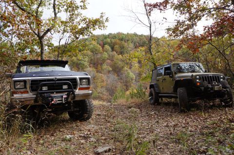 1979 Ford Bronco - Two off-road vehicles, a 1979 Ford Bronco and a Jeep Wrangler, rest on a leaf-covered trail during the fall.