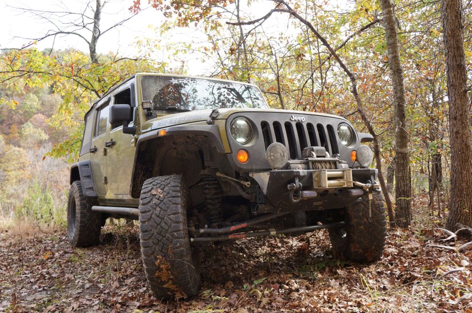 Olive green Jeep Wrangler with winch and off-road tires parked on a leaf-covered trail in the woods