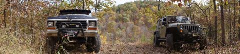 1979 Ford Bronco - Two off-road vehicles, a 1979 Ford Bronco and a Jeep Wrangler, are parked on a dirt trail surrounded by trees with fall foliage.