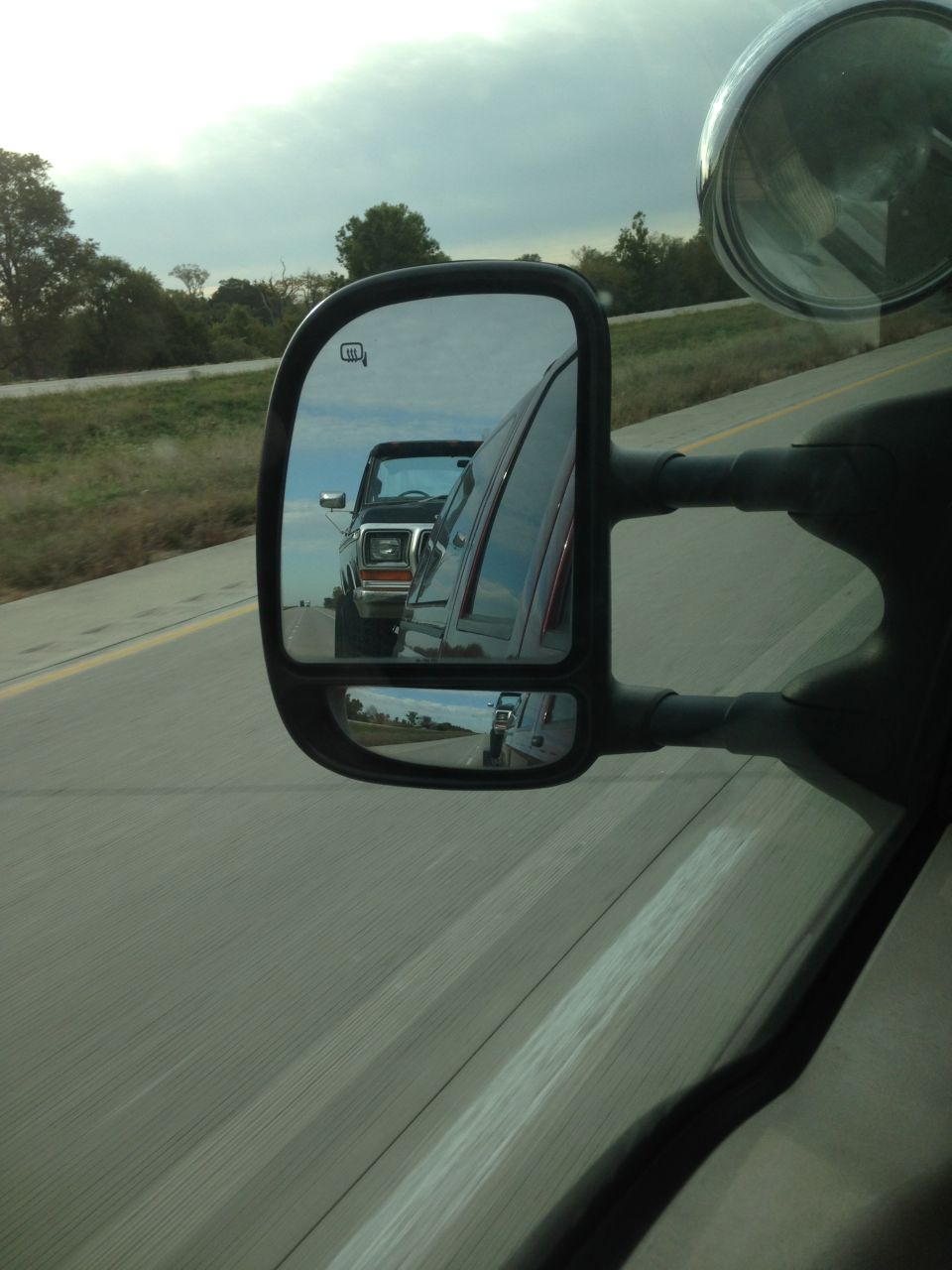 Side mirror view of a 1978 Ford Bronco driving on the highway. Another vehicle is visible in the distance