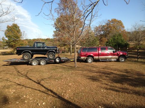 1979 Ford Bronco - A black pickup truck is loaded onto a trailer, with a red pickup truck parked nearby in a grassy, wooded area.
