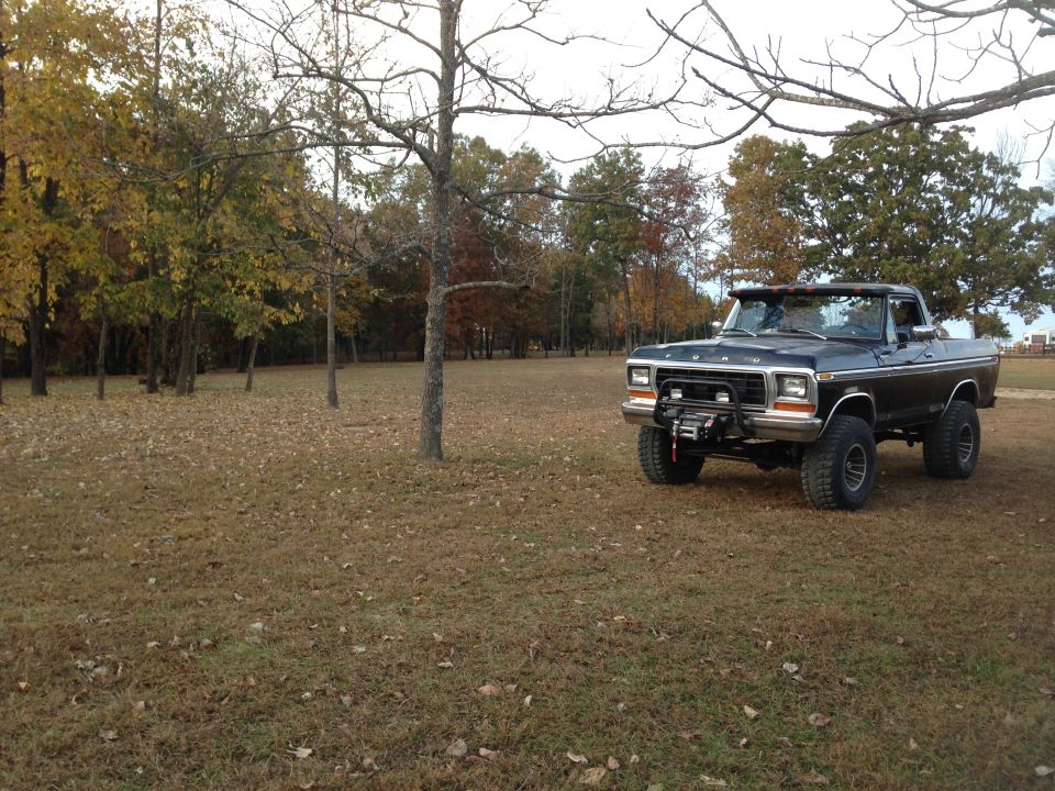 Black 1979 Ford Bronco with a winch on the front bumper, parked on a grassy field with fall foliage