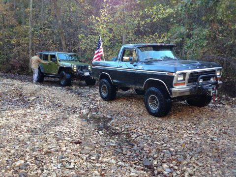 1979 Ford Bronco - Two off-road vehicles, a 1979 Ford Bronco and a Jeep Wrangler, are parked on a gravel path surrounded by trees.