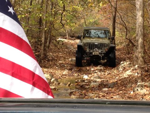 1979 Ford Bronco - An American flag partially obscures a Jeep Wrangler navigating a wooded trail during autumn.