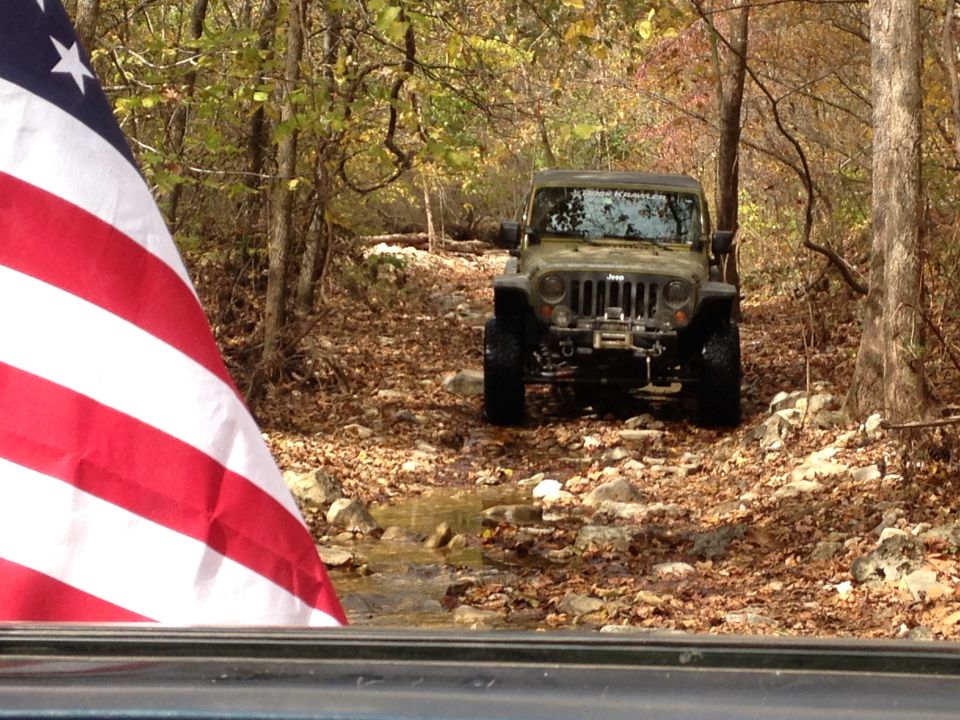 Olive green Jeep Wrangler on a rocky, leaf-strewn path with an American flag in the foreground
