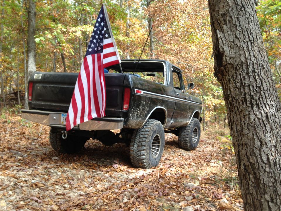 Black 1978 Ford Bronco with an American flag on the tailgate, seen from the rear in a wooded area
