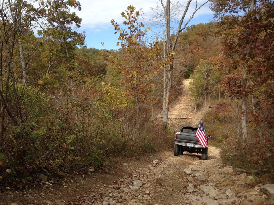 Pickup truck with American flag driving up a rocky dirt trail in autumn woods