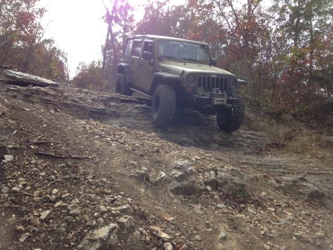 1979 Ford Bronco - Jeep Wrangler navigates a challenging off-road course with rocky obstacles and dense woods.
