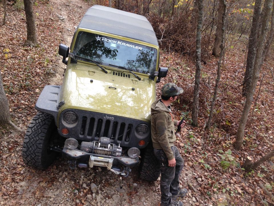 Yellow Jeep Wrangler parked on a wooded trail with a person leaning against it. Decal on windshield reads 'Rock Crawler'