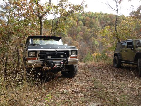 1979 Ford Bronco - A black Ford Bronco is parked on a dirt path, with another vehicle visible to the right, surrounded by autumn trees.