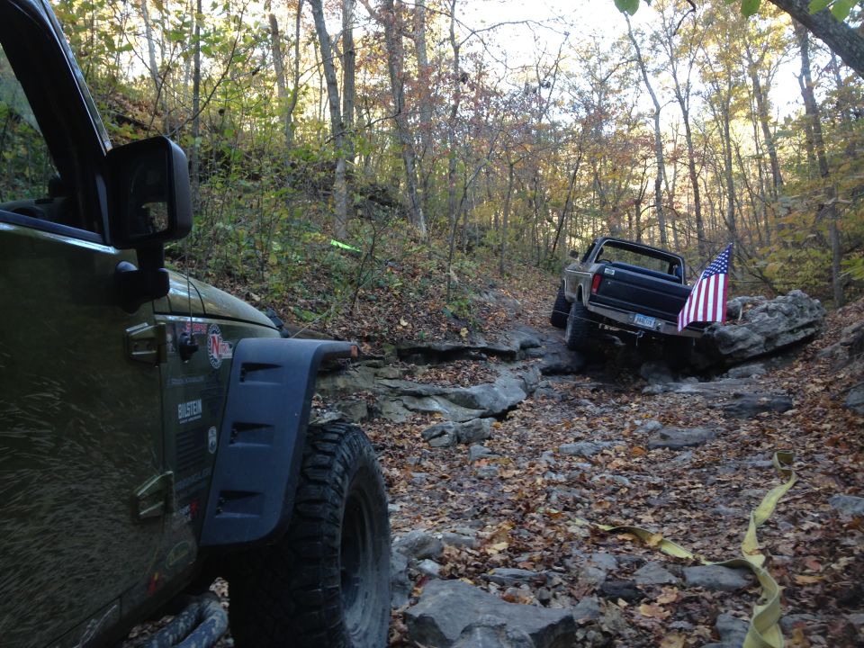 1979 Ford Bronco with American flag on rear, navigating a rocky trail behind a green Jeep