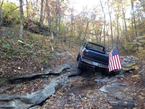 1979 Ford Bronco - A Ford pickup truck drives over rocks on a wooded trail, decorated with an American flag.