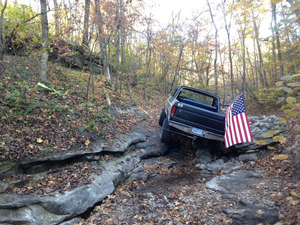 Black Ford pickup truck with American flag on rear, navigating rocky terrain in woods