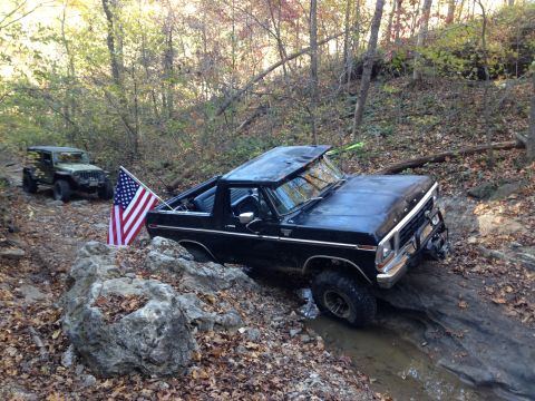 1979 Ford Bronco - A black 1979 Ford Bronco is navigating a rocky, wooded trail with an American flag mounted on the back. Another green Jeep is visible furth…