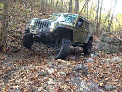1979 Ford Bronco - A green Jeep Wrangler is navigating a rocky, wooded trail covered in fallen autumn leaves. The vehicle has large off-road tires and a winch…