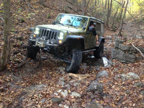 1979 Ford Bronco - A green Jeep Wrangler is navigating a rocky, wooded trail covered in fallen leaves. The driver is visible through the windshield, wearing a…