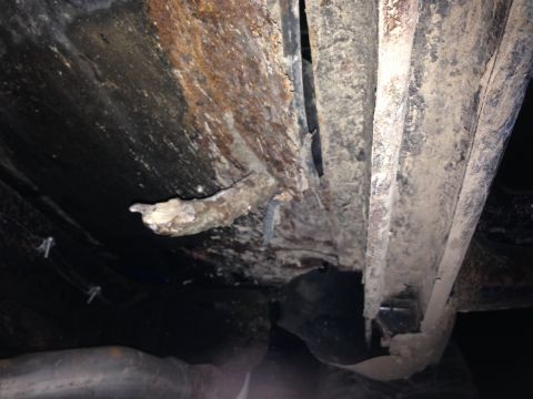 1979 Ford Bronco - The underside of a vehicle shows significant rust and corrosion on the frame and body panels. A broken piece of metal hangs from the frame,…
