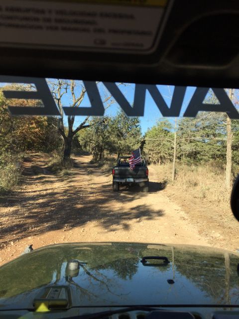 1979 Ford Bronco - View from inside a green Jeep looking at a dirt road with a black Ford pickup truck driving ahead, adorned with an American flag.