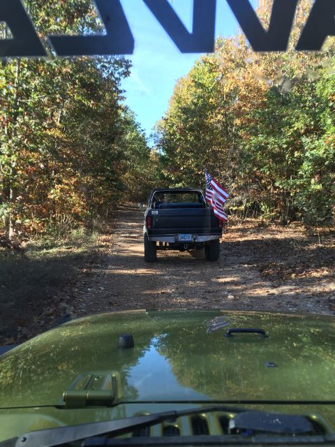 1979 Ford Bronco - A black Ford pickup truck with an American flag in the bed is driving down a dirt road lined with autumn trees. The photo is taken from the…
