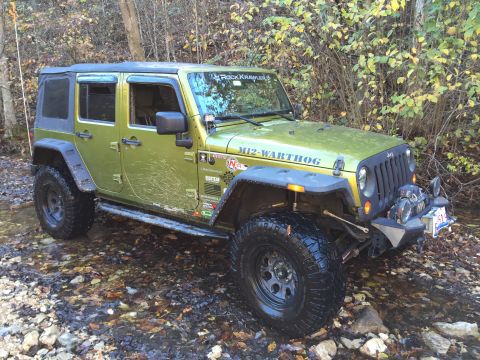 1979 Ford Bronco - A bright green Jeep Wrangler is shown from a 3/4 front angle, driving through a shallow stream with fallen leaves on the ground. The Jeep h…