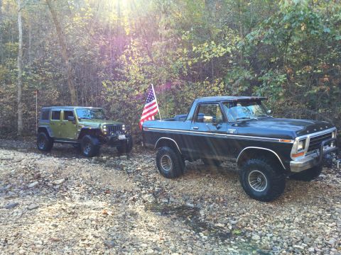 1979 Ford Bronco - A black 1979 Ford Bronco with an American flag attached to its antenna is parked on a rocky surface next to a green Jeep Wrangler. The vehi…