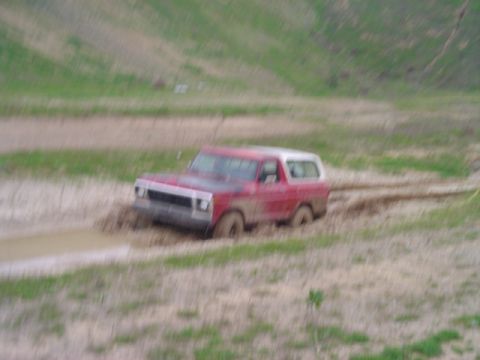 1978 Ford Bronco - A red 1978 Ford Bronco navigates a muddy off-road course, with its front-left wheel deep in the mire.
