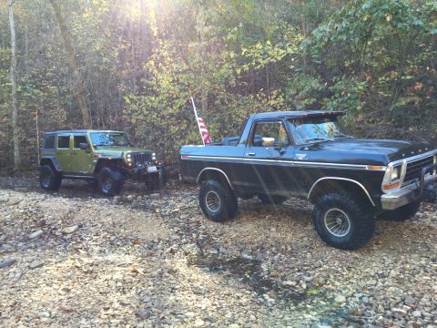 1979 Ford Bronco - A black Ford Bronco with an American flag attached to the antenna is parked on a rocky stream bed next to a green Jeep Wrangler.