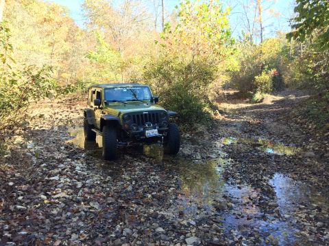 1979 Ford Bronco - A green Jeep Wrangler is driving through a shallow stream bed on a wooded trail during autumn. The vehicle is angled towards the camera, sh…