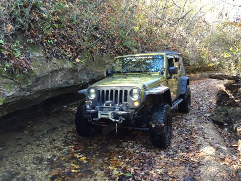 1979 Ford Bronco - A green Jeep Wrangler is parked in a shallow stream bed surrounded by trees and rocks. The vehicle has a winch mounted on the front bumper…