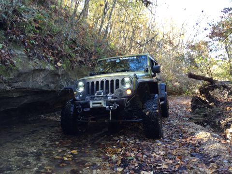 1979 Ford Bronco - A green Jeep Wrangler is driving through a shallow stream in a wooded area with fallen leaves on the ground. The Jeep has large off-road ti…