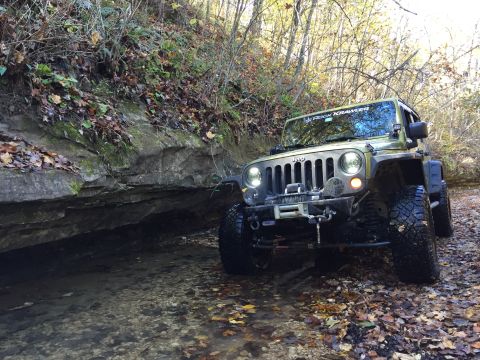 1979 Ford Bronco - A green Jeep Wrangler is parked in a shallow stream bed, surrounded by fallen autumn leaves and trees. The vehicle is equipped with off-roa…