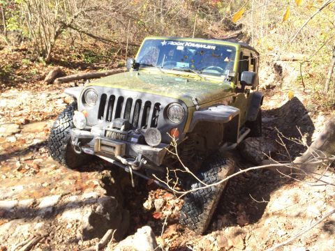1979 Ford Bronco - A green Jeep Wrangler is navigating a rocky, wooded trail with its front tires deep in a rut. The vehicle has a winch mounted on the front…