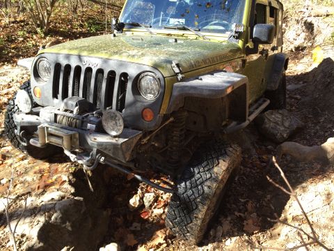 1979 Ford Bronco - A close-up, low-angle view of the front of a lifted, olive-green Jeep Wrangler off-roading on a rocky, wooded trail. The Jeep is equipped w…