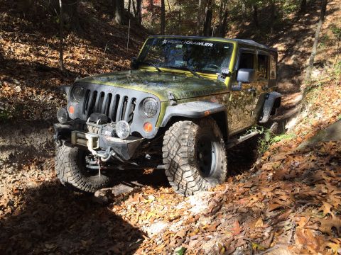 1979 Ford Bronco - A green Jeep Wrangler is shown from a 3/4 front angle, navigating a wooded trail covered in fallen autumn leaves. The vehicle has off-road…