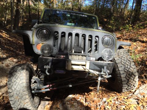 1979 Ford Bronco - A modified Jeep Wrangler is shown from a low-angle front 3/4 view, parked on a wooded trail covered in fallen leaves. The Jeep features lar…