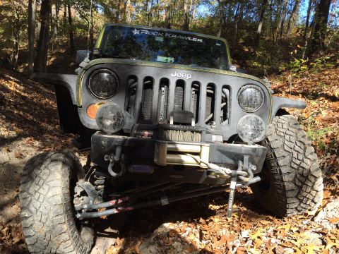 1979 Ford Bronco - A front 3/4 view of a modified Jeep Wrangler parked on a wooded trail with fallen autumn leaves on the ground. The vehicle features large o…