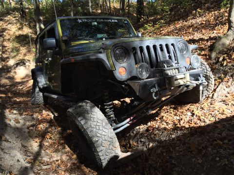 1979 Ford Bronco - A lifted, olive green Jeep Wrangler is shown on a dirt trail covered in fallen leaves. The vehicle has a winch mounted on the front bumper…