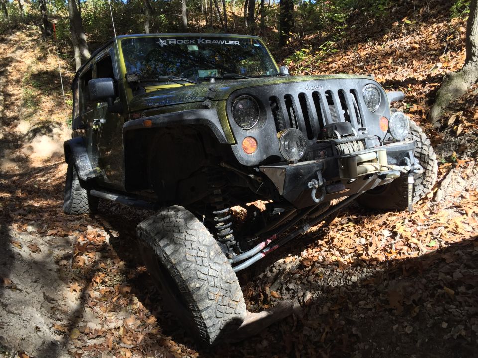 Lifted olive green Jeep Wrangler with winch on a leaf-covered dirt trail.