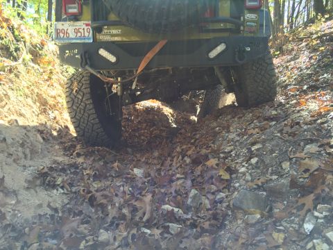 1979 Ford Bronco - The rear of a green Jeep Wrangler is shown from a low angle, driving on a leaf-covered, rocky trail. The vehicle has an aftermarket bumper…