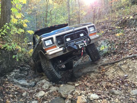 1979 Ford Bronco - A black 1979 Ford Bronco is navigating a rocky, wooded trail with fallen leaves on the ground. The front of the vehicle is angled upwards,…