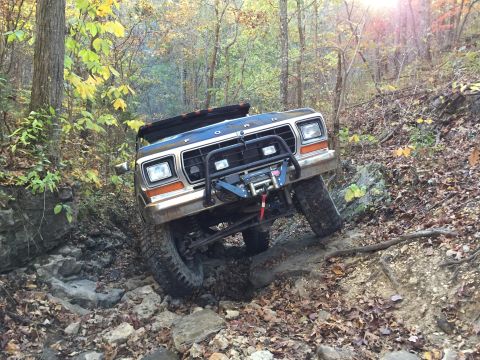 1979 Ford Bronco - A dark colored Ford Bronco is navigating a rocky, wooded trail in the fall. The vehicle is angled sharply to the right, with its front tire…