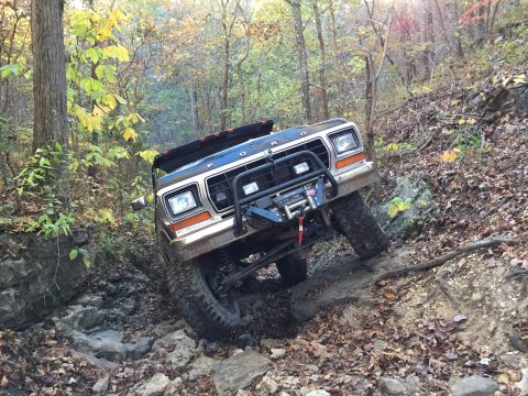 1979 Ford Bronco - A dark colored Ford Bronco is navigating a rocky, wooded trail during the fall. The vehicle is tilted at an angle as it climbs over large r…
