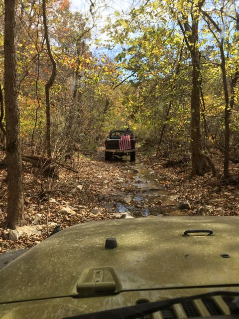 1979 Ford Bronco - A dark-colored pickup truck with an American flag attached to the tailgate drives down a rocky, wooded stream bed during autumn. The image…