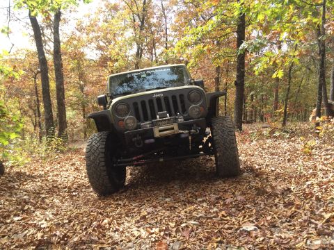 1979 Ford Bronco - A tan Jeep Wrangler is parked on a leaf-covered forest floor, surrounded by trees with autumn foliage. The vehicle features large off-road…