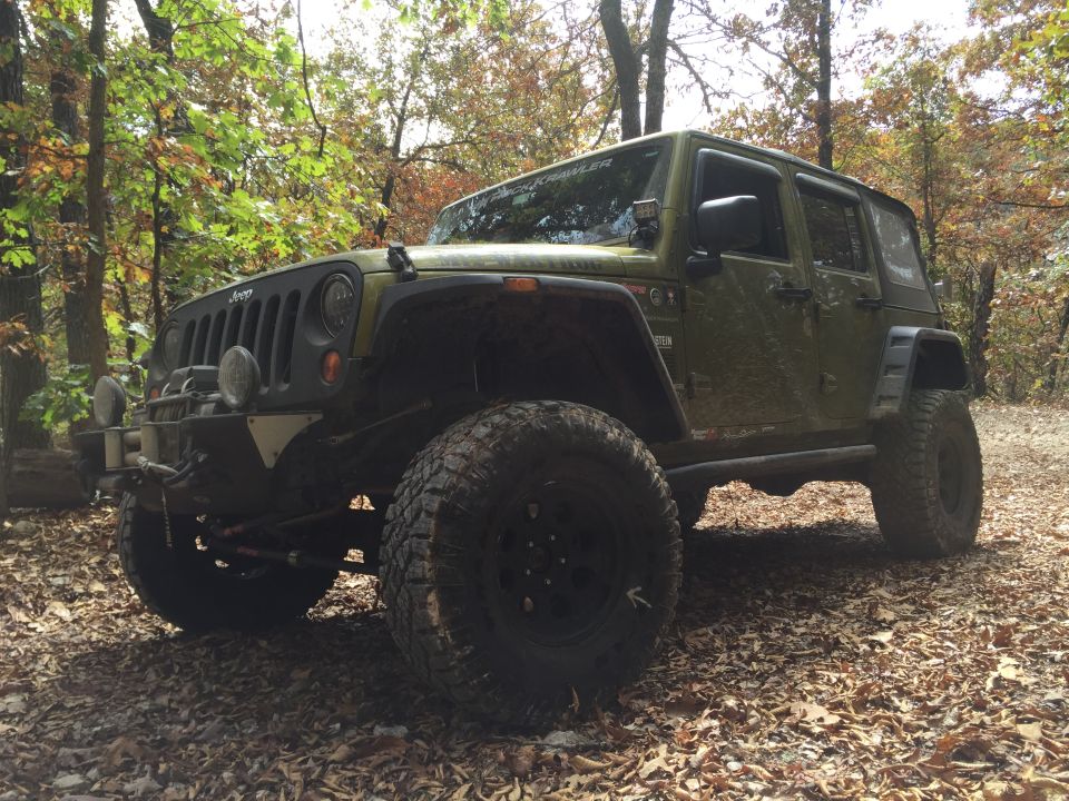 Green Jeep Wrangler with large tires and winch parked on a leaf-covered path in a forest.