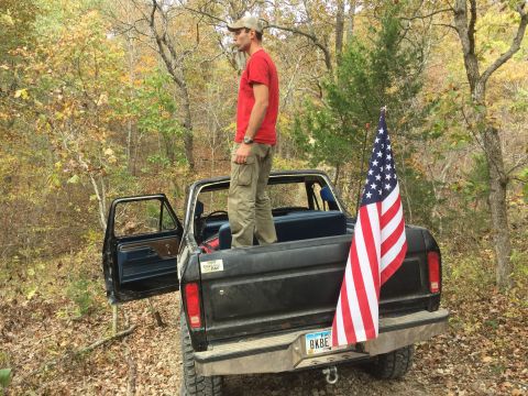 1979 Ford Bronco - A man stands in the bed of a black pickup truck with an American flag attached to the tailgate. The truck is parked on a dirt road surround…
